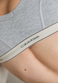 Šviesiai pilkas medvilninis liemenėlė su apvaliu iškirpimu ir plačia elastinga juostele su juodu „Calvin Klein“ logotipu ant kreminės spalvos fono.