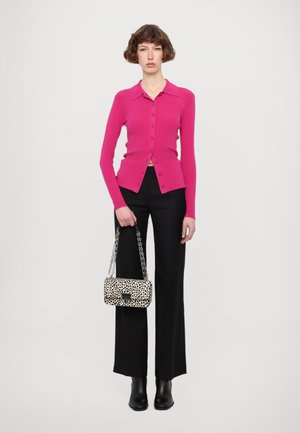 Magenta knitted cardigan with a collar and buttons, paired with black trousers. Holding a cream and black spotted handbag with a chain strap.
