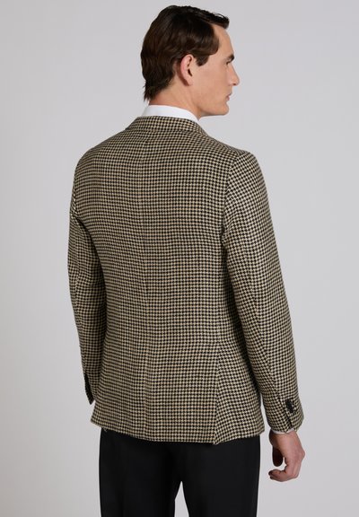 Mann wird seitlich gezeigt, trägt einen braun-schwarzen Hahnentritt-Blazer mit weißem Hemdkragen und schwarze Hose vor grauem Hintergrund.