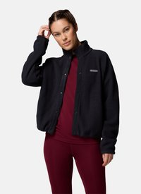 Jonge vrouw die haar haar goed doet, draagt een zwarte Columbia fleecejas en bordeauxrode sportlegging tegen een effen achtergrond.