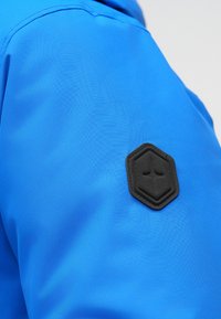 Blue jacket with a smooth texture, featuring a black hexagonal logo patch on the upper arm. The fabric appears lightweight and durable.