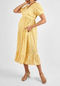 Schwangere Frau in einem gelben, floralen Midikleid mit Puffärmeln und goldenen High Heels, die mit einer Hand auf der Hüfte und mit der anderen am Hals posiert.