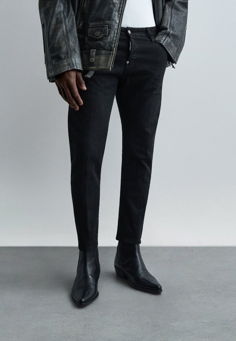 Homme portant une veste en cuir noire, un t-shirt blanc, un pantalon noir slim et des bottines noires à bout pointu, debout sur un sol gris.