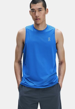 Homme portant un débardeur bleu vif et un short sombre, debout les mains dans les poches devant un fond uni.