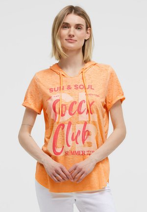 Woman with short blonde hair wearing an orange short-sleeve hoodie with pink text, standing with hands loosely clasped in front.
