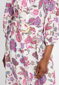 Weißes Blumenkleid mit pinken, blauen und grünen Mustern. Es hat eine Knopfleiste und einen breiten Gürtel mit einer runden, naturfarbenen Schnalle.