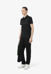 Polo negro de manga corta, con una franja verde contrastante a través del pecho, combinado con pantalones negros y zapatillas blancas.