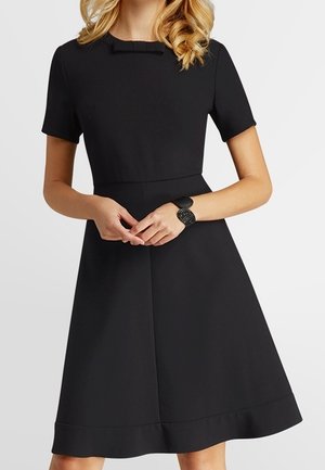 Femme portant une robe noire évasée à manches courtes avec un petit nœud au niveau de l'encolure et un bracelet noir texturé.