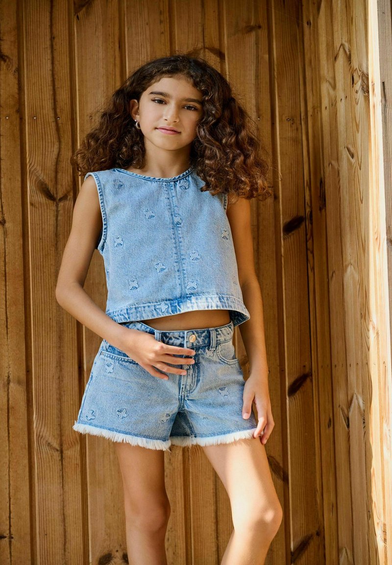 Denimcrop topp med broderade mönster, i kombination med shorts med fransar. Ljusblå färg, enkel design, avslappnad passform och ärmlös.