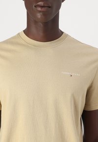 Khaki cotton t-shirt with a crew neckline, featuring a white "TOMMY JEANS" logo on the left chest. Simple, smooth texture.