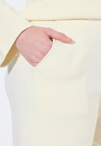 Cremfarbener Strickartikel mit einer Seitentasche und einem texturierten, weichen Stoff. Das Design umfasst gerippte Bündchen und eine eng anliegende Passform.