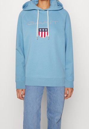 Kobieta w jasnoniebieskiej bluzie z kapturem z logo GANT i herbem, nosząca jasnoniebieskie dżinsy, stojąca na jednolitym tle.