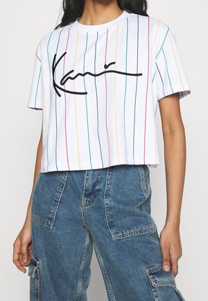 Kobieta ubrana w biały krótki T-shirt z wielokolorowymi pionowymi paskami i czarnym logo w stylu pisma, połączony z niebieskimi dżinsowymi spodniami typu cargo.