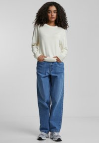 Pull crème clair avec des poignets côtelés, associé à un jean en denim bleu ample. Le modèle porte des baskets grises. Tenue simple et décontractée.