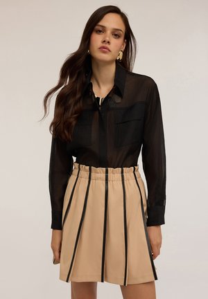 Schwarzes, durchscheinendes Langarmshirt mit zwei Brusttaschen, kombiniert mit einem beigen Rock mit senkrechten schwarzen Streifen und elastischem Bund.