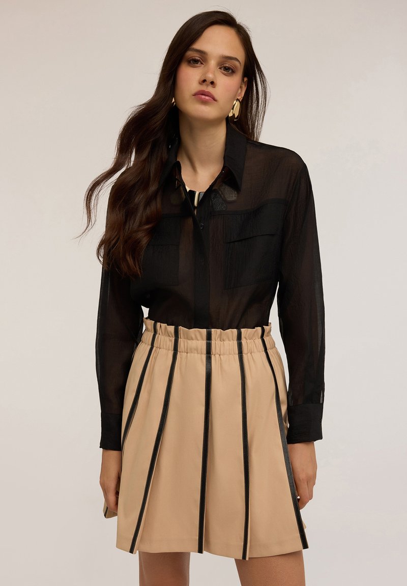 Camicia nera trasparente a maniche lunghe con due tasche sul petto, abbinata a una gonna beige con strisce nere verticali e una vita elasticizzata.