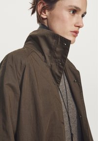 Zielona oliwkowa kurtka z wysokim kołnierzem, o gładkiej fakturze i guzikowych akcentach, nałożona na szary sweter o drobnym wzorze dzianiny.