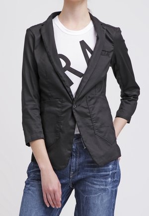 Personne portant un blazer noir à un bouton par-dessus un t-shirt blanc avec un motif graphique et un jean bleu déchiré, avec une main dans la poche.