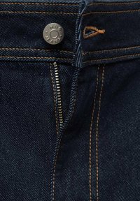 Donkerblauwe denim jeans met contrasterende oranje stiksels, een metalen knoop met het label "MNG JEANS" en een zilveren ritssluiting. Textuur is glad met een lichte glans.