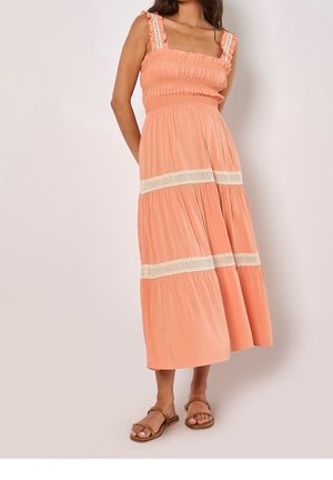 Femme portant une robe maxi pêche sans manches avec bordure en dentelle et bretelles à volants, assortie à des sandales plates marron, debout devant un fond uni.