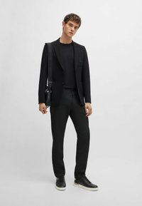 Blazer noir avec deux poches avant, associé à un t-shirt noir ajusté et un pantalon texturé sombre. Des baskets noires et un sac bandoulière complètent le look.