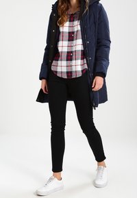 Marinblå vadderad jacka med huood, röd och svart rutad skjorta, svarta slim fit-jeans, vita sneakers, uppställd mot en neutral bakgrund.