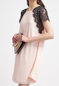 Light pink dress with black lace short sleeves, curved hem, and subtle drape; paired with a black and beige clutch bag.