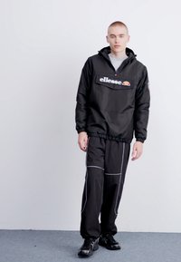 Giacca a vento nera con zip a metà, caratterizzata da un logo sul petto e polsini elasticizzati. Abbinata a pantaloni neri con profilature bianche.