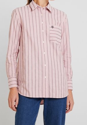 Woman wearing a light pink button-up shirt with thin vertical stripes and a front pocket, paired with blue jeans.