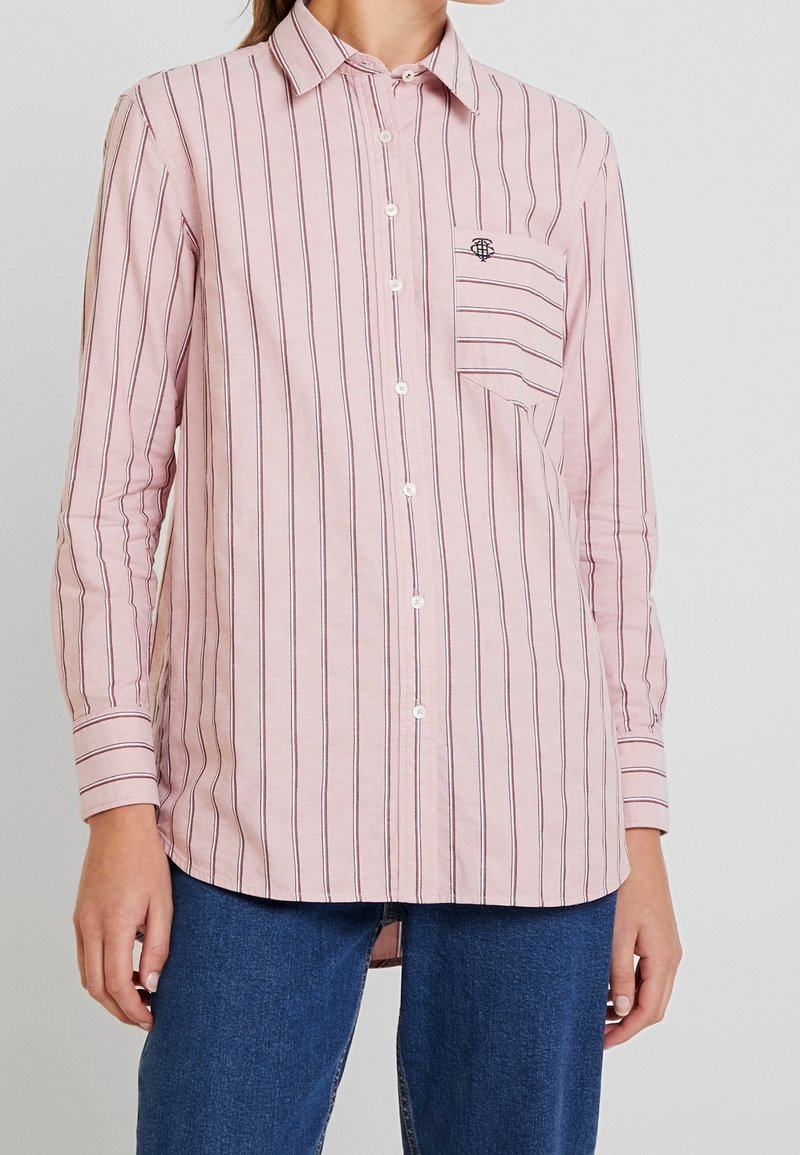 Femme portant une chemise rose pâle à fines rayures verticales avec une poche avant, assortie d'un jean bleu.