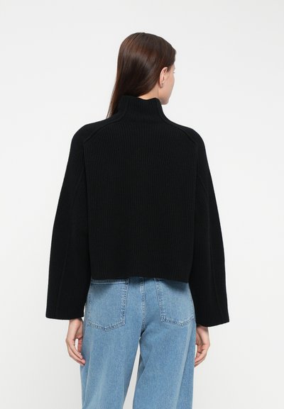 Black ribbed sweater with a high collar and cropped design. Features wide sleeves and a textured finish, paired with light blue jeans.