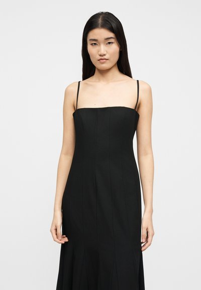 Sieviete ar gariem taisniem melniem matiem, valkājot bezrokainu melnu kleitu ar plāniem lencītēm uz gaiša, vienveidīga fona.