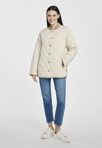 Jasno-beżowa pikowana kurtka z zatrzaskami, kieszeniami bocznymi i okrągłym dekoltem. Noszona na niebieskie dżinsy i białe trampki.