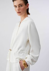Jeune femme portant un sweat à capuche blanc zippé avec taille à cordon, pantalon blanc, boucles d'oreilles créoles dorées et trois bracelets dorés au poignet.