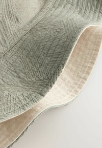 Cappello a secchiello verde chiaro con cuciture geometriche testurizzate e fodera interna color crema su sfondo bianco.