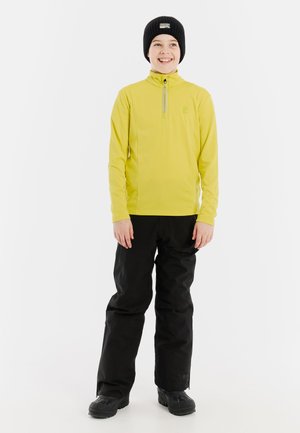 Geel, langemouwen shirt met een kwart rits, gecombineerd met zwarte sneeuwbroeken. Een zwarte gebreide muts maakt de outfit compleet. Glad textuur overal.