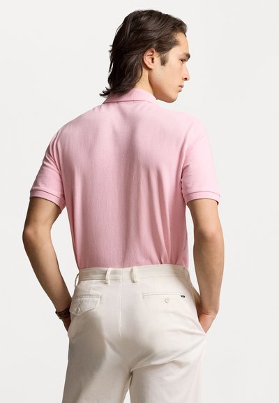 Tricou polo de culoare roz deschis, din material texturat, cu mânecă scurtă și guler. Purtați-l cu pantaloni de culoare crem, arătând din spate.