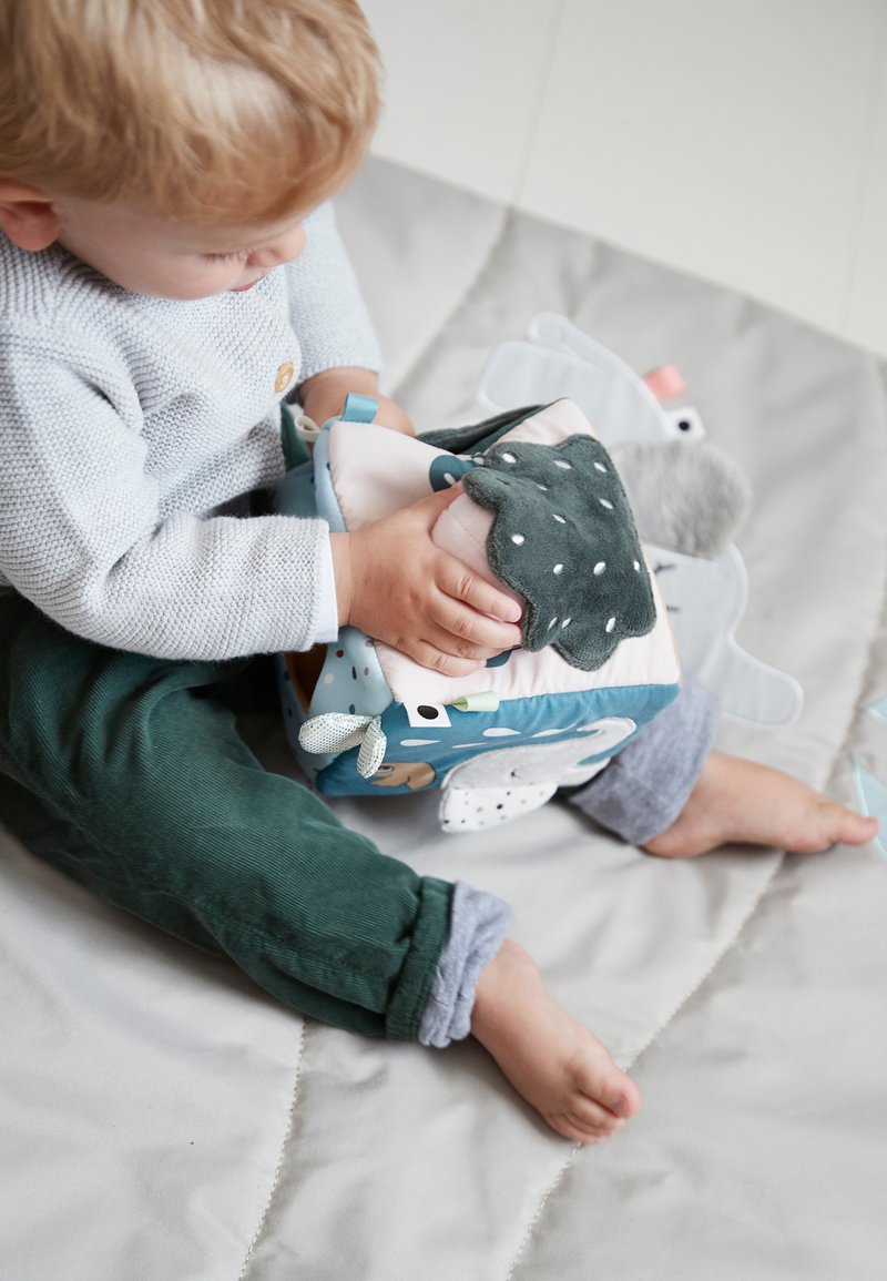Soft fabric activity cube with various textures, colors (blue, green, beige), and patterns. Child holds a plush, polka-dotted flap.