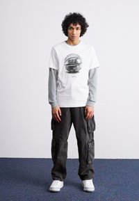 Camiseta blanca con un gráfico negro, superpuesta sobre una camiseta de manga larga gris. Pantalones cargo oscuros y zapatillas blancas completan el conjunto.
