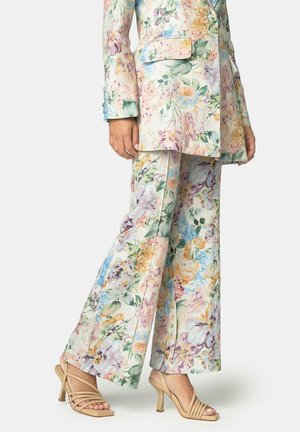 Mujer vistiendo un blazer de flores en tonos pastel y pantalones anchos a juego, con sandalias de tacón altas beige con tiras, de pie contra un fondo liso.