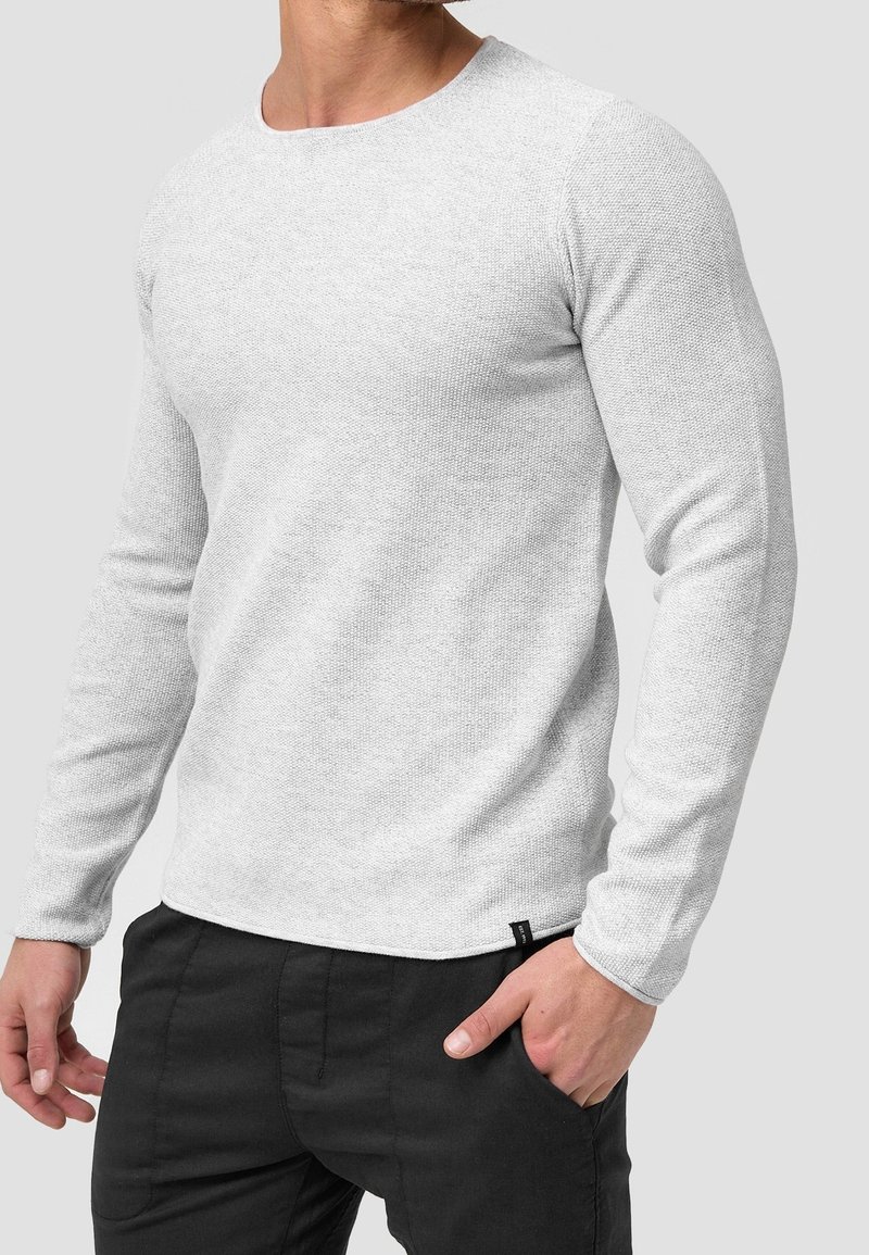 Homme portant un pull à manches longues texturé gris clair et un pantalon noir, une main dans la poche, sur un fond clair uni.