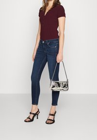 Mujer vestida con una blusa burdeos de botones y jeans ajustados oscuros, sosteniendo un bolso metálico plateado y llevando sandalias de tacón negras.