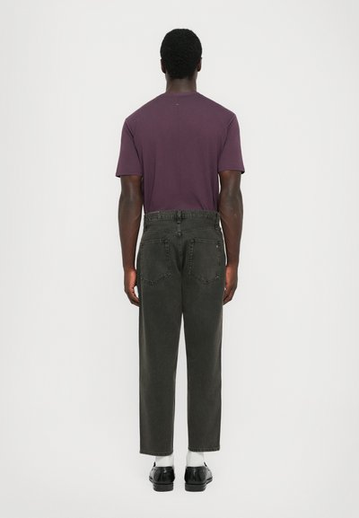 Man som står med ryggen mot kameran och bär en lila t-shirt, mörkgrå högmidjade jeans, vita strumpor och svarta skor mot en vit bakgrund.