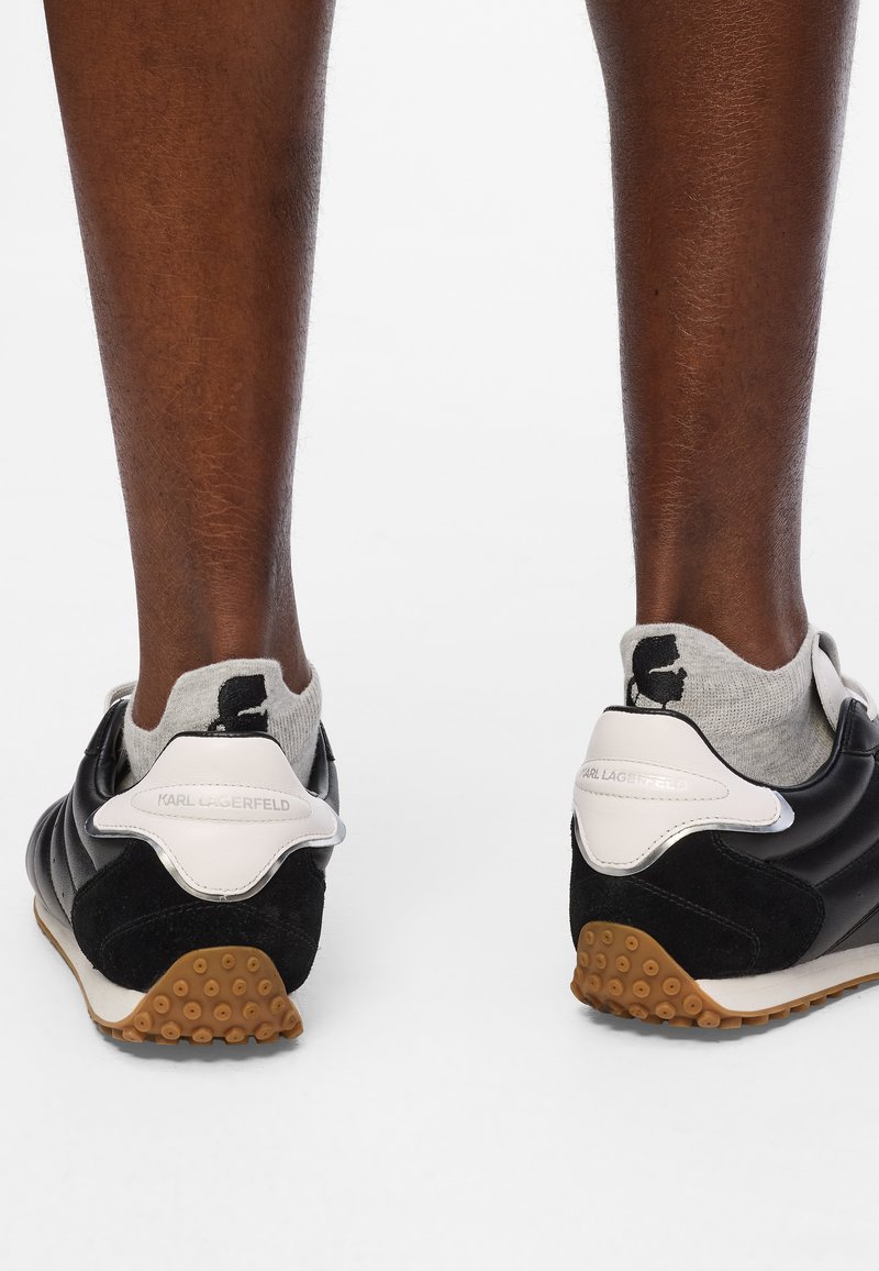 Zapatillas deportivas en negro y gris con materiales de ante y cuero, que presentan una suela de goma con dibujos circulares y un acento en blanco en el talón.