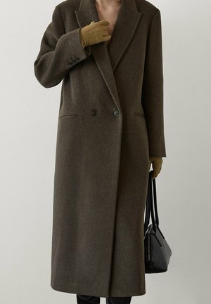 Woman wearing a long brown wool coat, tan gloves, holding a small black handbag against a plain background.