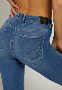 Jeans de denim de lavado oscuro con un diseño ajustado, que presentan un detalle de bolsillo único con embellishments metálicos. Etiqueta negra de "GUESS" en la cintura.