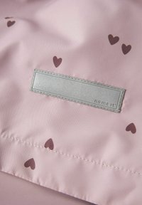 Tissu rose clair avec de petits cœurs foncés imprimés, doté d'une zone rectangulaire cousue pour une étiquette réfléchissante.