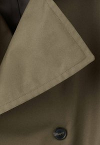 Tela de color verde oliva con una textura suave, que presenta una solapa grande y un detalle de botón negro. Las costuras son limpias y bien acabadas.