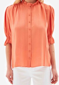 Blouse couleur pêche à rayures verticales, col volanté et devant boutonné. Manches bouffantes avec poignets élastiques.