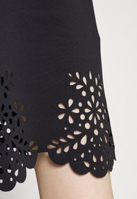 Svart tygkjol med scallopad nederkant, med utskurna blommönster för textur och detaljer. Närbild av den nedre delen.